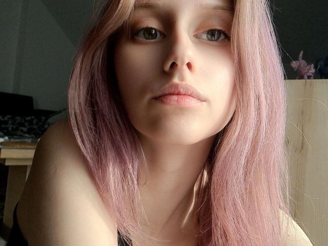 LunaXKiss with pale pink hair, green eyes, and pink lips, looking straight at the camera with a neutral expression, wearing a black top, showing her bare shoulders.