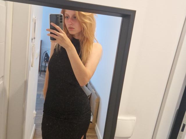LunaXKiss, blonde, in black sleeveless dress, holding phone, mirror selfie, looking serious, slim, small tits, fair skin, reflection, wooden floor, white wall, door, fan in background.