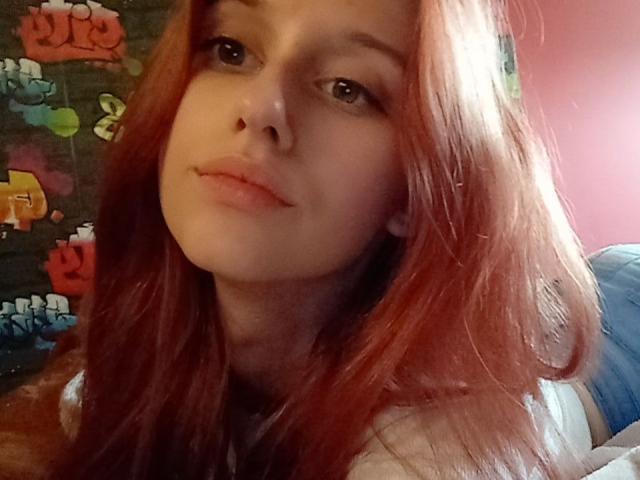 LunaXKiss has pale skin, red hair, and a slight pout with pink lips. She's wearing a white shirt, looking at the camera with a relaxed expression.