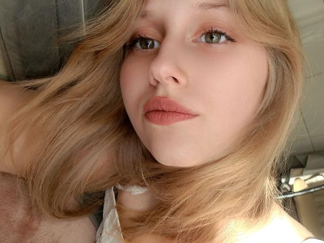LunaXKiss has light blonde hair, green eyes, and pinkish lipstick. Her soft face is close-up, showing her slight smile and fair skin. She's wearing a white top.