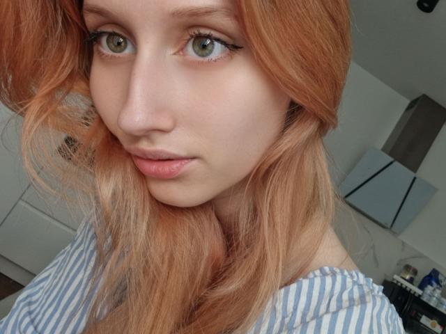LunaXKiss has pale skin, green eyes, and red-orange hair, wearing a blue-striped shirt, with a slight pout on her pink lips, in a soft light selfie.