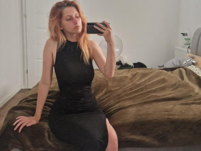 LunaXKiss with red hair, wearing a black, sleeveless, high-neck dress, sitting on a brown, fuzzy bed, taking a mirror selfie, showing her ass and tits.