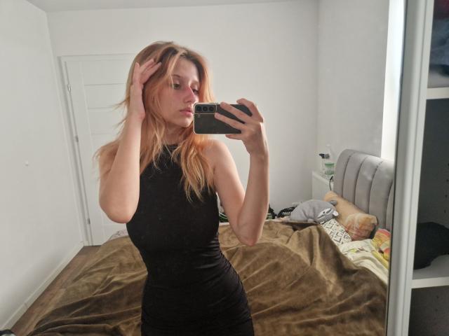 LunaXKiss with blonde hair, wearing a tight black dress, holding a phone, taking a mirror selfie, showing her slim body and small tits, in a bedroom.
