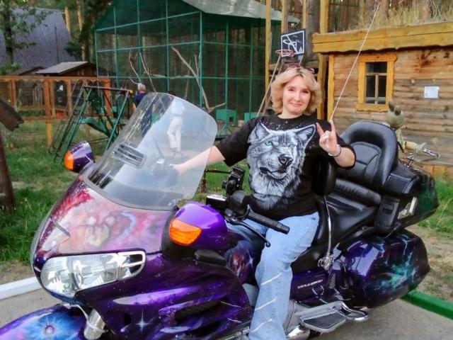 VAlica with blonde hair, wearing a black wolf shirt and blue jeans, sits on a purple, star-patterned motorcycle, making a rock-and-roll hand sign, smiling.