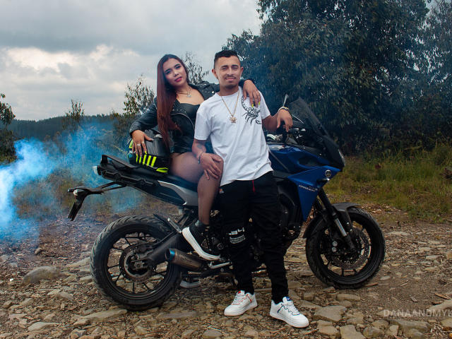 DannaAndMikes, con los senos y el trasero visibles, está sentada en una motocicleta azul, vistiendo una chaqueta negra y pantalones cortos, mientras que Mikes, con una camiseta blanca y pantalones negros, está de pie frente a ella..