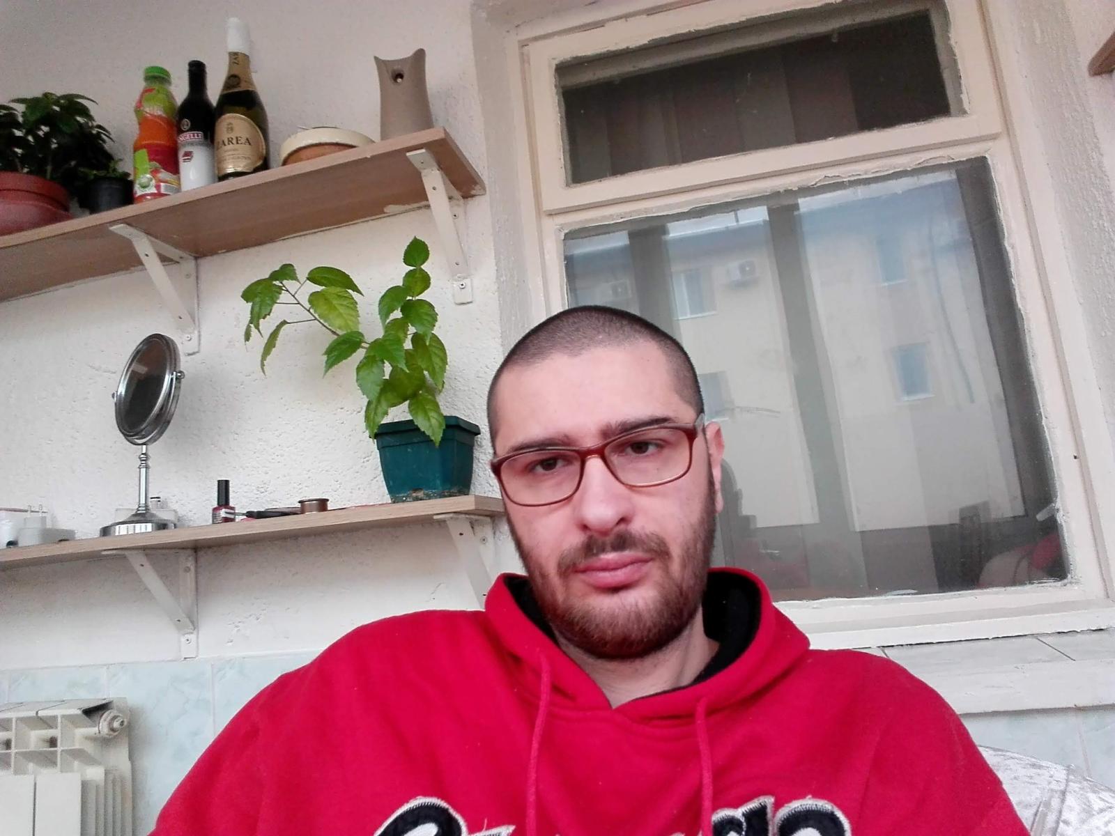 BigBoby with a shaved head, red glasses, and a red hoodie, has a beard and is looking at the camera, sitting in a room with a window.