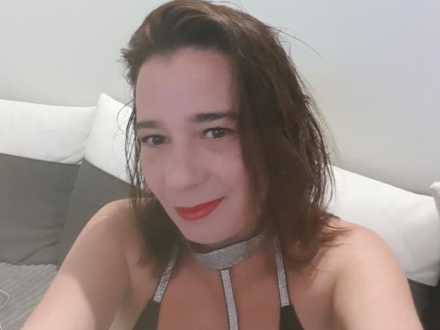 FloryanCouple has medium brown hair, red lipstick, and a silver choker with straps, showing a slight smile, sitting on a bed with white and gray pillows.