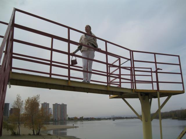 LoveFM-hot with white pants and tan jacket stands on a red metal bridge, holding a brown bag, looking down. Overcast sky, river, and autumn trees in background.