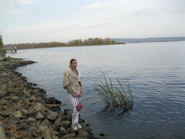 LoveFM-hot with blonde hair, wearing a beige shawl, white pants, and red purse, stands on rocky shore by calm water, near tall grass.