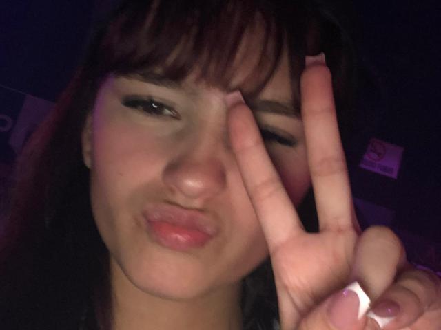 MelodyMansson's pouty face with dark brown hair and bangs, making a "V" sign with her right hand, has pinkish-purple nail polish and slightly parted pink lips.