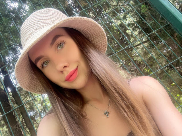 SannyManny with long brown hair, green eyes, pink lips, wearing a straw hat and cross necklace, showing off her tits in a forest selfie.