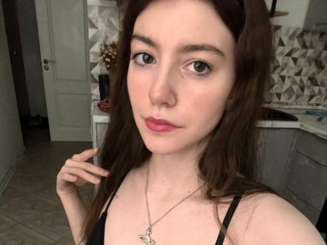VinnaSkyHott has pale skin, green eyes, and pink lipstick, with brown hair and a black top, holding a lock of hair, wearing a star necklace.