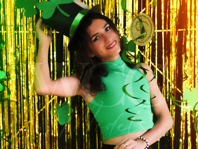 AndyDupont with curly black hair, green top, and black hat, smiling, holding green and gold party decorations, wearing a green and gold bracelet, against shiny gold and green background.