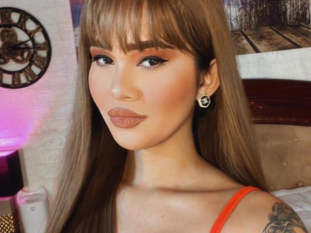 LoveMeKara with long brown hair, bangs, and red top, showing off tattoos on her right shoulder, wearing black floral earrings, and pouty lips. Pink light in background.