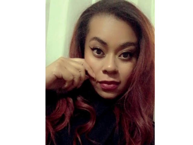 Mya Spencer, con cabello rojo degradado, un cuello alto negro, y delineador de ojos alado, con el mentón apoyado en la mano, mirando a la cámara, labios carnosos, piel marrón media, y maquillaje sutil..