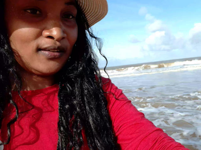 NastyGee mit dunklen, lockigen Haaren, trägt ein rotes Langarmshirt und einen Strohhut und steht am Meer, im Hintergrund ist ein blauer Himmel mit weißen Wolken zu sehen..