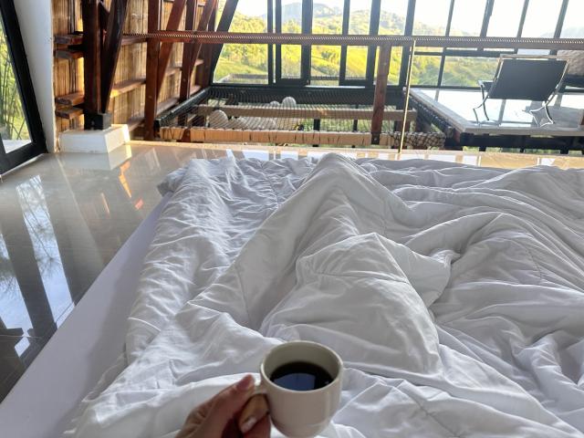 La main de Leared tient une tasse blanche de café noir, posée sur un lit désordonné avec des draps blancs, dans une pièce moderne en bois, baignée de lumière du soleil..