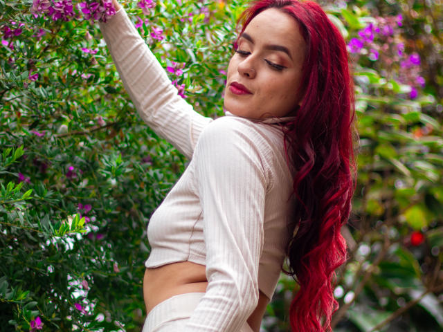 MaiaPark with red hair, wearing a white ribbed crop top, reaching for pink flowers, showing her tits and a slight belly, smiling with closed eyes in a garden.