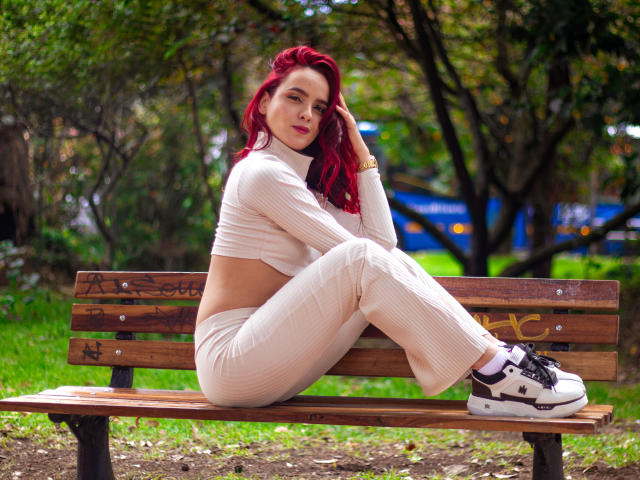 MaiaPark with red hair, sitting on a wooden bench, wearing a cream ribbed crop top and pants, white sneakers, looking at the camera, hand in hair.