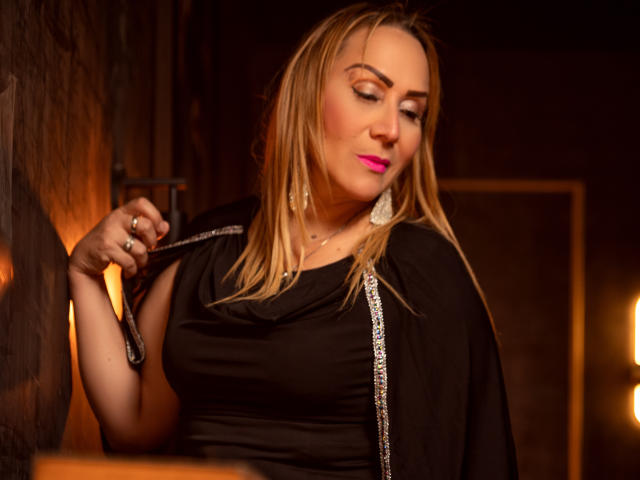 DianeOasis with blonde hair, pink lipstick, and black dress, holding a black whip, looking down with a slight smile, wearing large white earrings, in a dimly lit room.