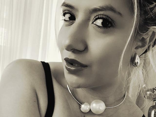 ValeriaHunter69 has a pretty face with shiny eyes, dark lipstick, and a pearl necklace. She's wearing a black strap, and her ear is adorned with a teardrop earring.