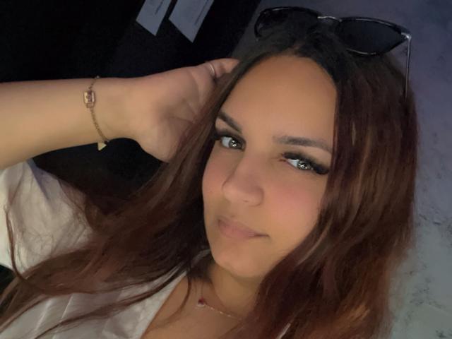 RedQueenX has long brown hair, green eyes, and a cute face. She's wearing black eyeliner, a white shirt, a bracelet, and a necklace with her hand in her hair.