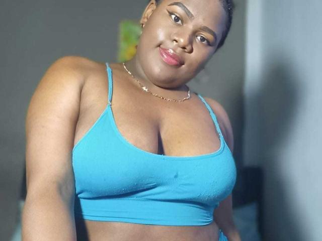 EbonySkyX with dark skin, blue sports bra, showing her big tits, slight smile, gold necklace, and glossy pink lips, looking at the camera, in a softly lit room.