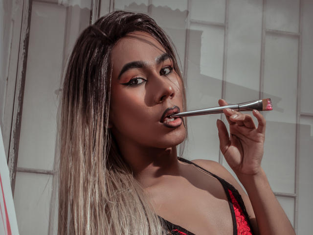 ScarlettHotSweet with long blonde hair, dark eyebrows, and red eyeliner, licking a silver lipstick brush with pink tip, wearing a black and red lace top.