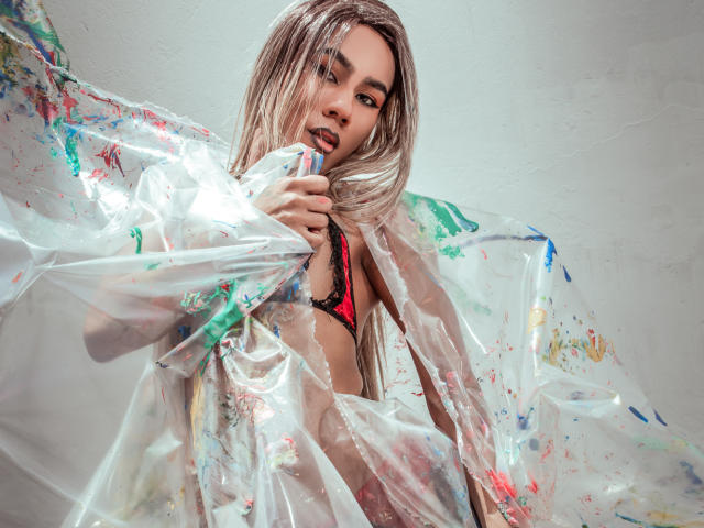 ScarlettHotSweet with long blonde hair, red lace bra, and transparent plastic sheet covered in colorful paint splatters, holding the sheet, looking at the camera with a slightly open mouth.