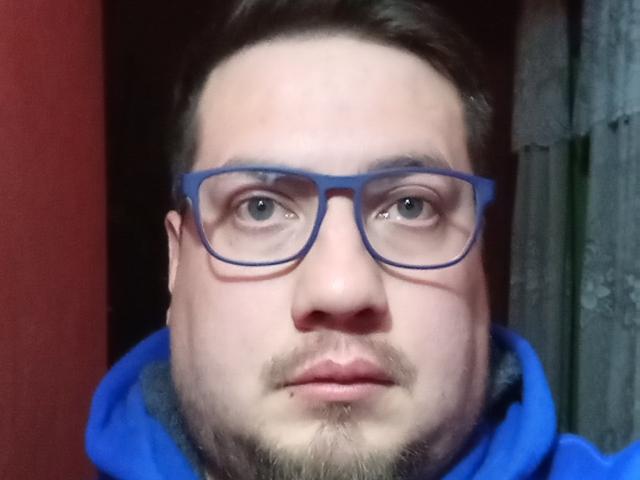 SnowBally has a chubby face, blue glasses, and a blue hoodie. He's got a beard, light skin, and short brown hair. His eyes are blue and he's looking straight at the camera.