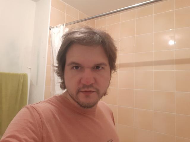 NectarineFad, a chubby guy with brown wavy hair and a beard, wearing a pinkish t-shirt, in a bathroom with orange tiles and a green towel.