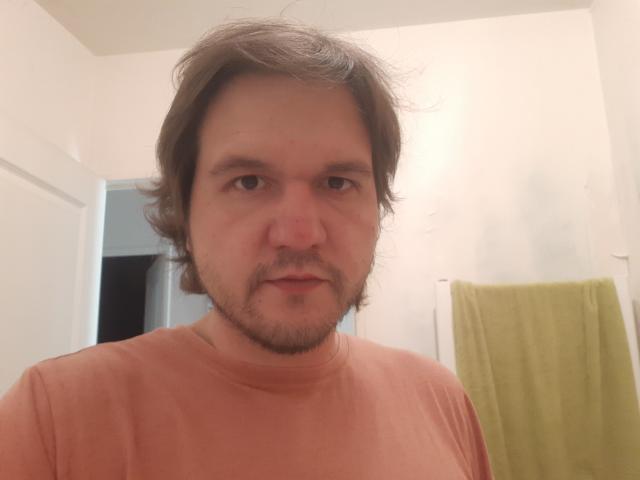 NectarineFad, a chubby guy with brown wavy hair, a light beard, and a serious expression, wearing a pinkish-orange t-shirt, standing in a bathroom with green and yellow towels.