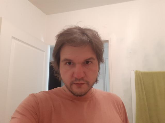 NectarineFad has messy brown hair, a beard, and a serious expression, wearing a pinkish-orange shirt, standing in a bathroom with a green towel on the right.