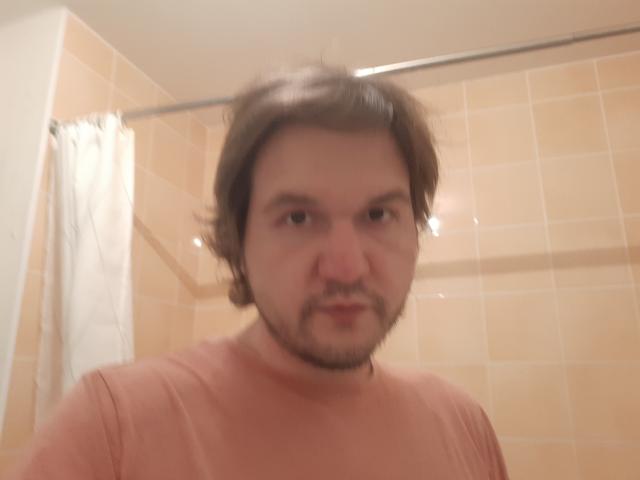 NectarineFad has a slightly blurry, average-looking face with brown, wavy hair and a beard. He's wearing a pinkish-red shirt. Bathroom with tan tiles in the background.