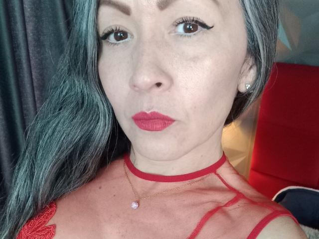 NicoletteShea has gray hair, red lipstick, and wears a red sheer top with a choker. She has eyeliner, a pink nose, and a necklace. Her tits are visible.