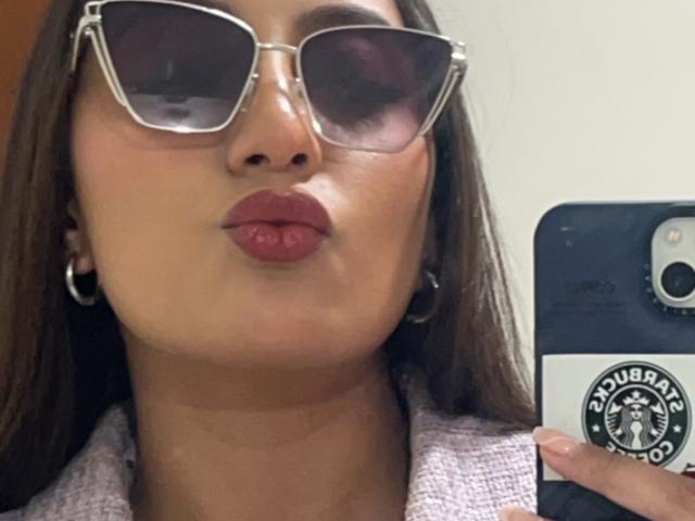 DianaFernandezz with pouty red lips, brown hair, wearing silver-framed sunglasses and a pinkish-gray knitted sweater, holding a phone with a Burberry sticker. Her hand is on the right.