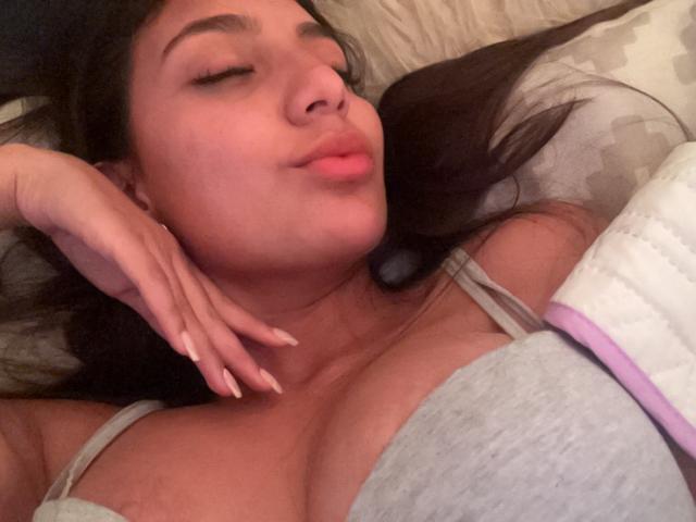 DianaFernandezz with her eyes closed, pouty lips, and long dark hair, lying on a bed. She's wearing a gray top, exposing her large boobs, with manicured nails.