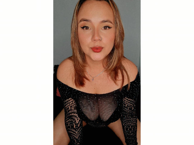 Natalie Rey, con i capelli castano chiaro, indossa una maglia nera trasparente in pizzo che mette in mostra il suo seno piccolo, e una collana d'argento, con le labbra rosse che fanno un labbro..