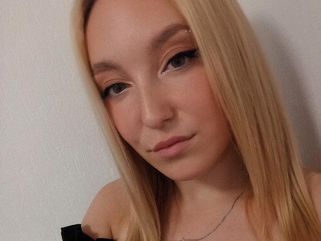 DinaToni ha i capelli biondi, un trucco leggero e un sorriso appena accennato, con gli occhi rivolti in avanti e un piccolo piercing al naso. Le sue tette non sono visibili nell'immagine..