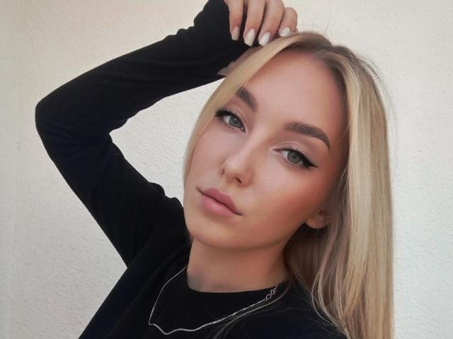 DinaToni has blonde hair, green eyes, and wears a black long-sleeve shirt with a thin necklace. She's holding her hair up, showing off her white nail polish.