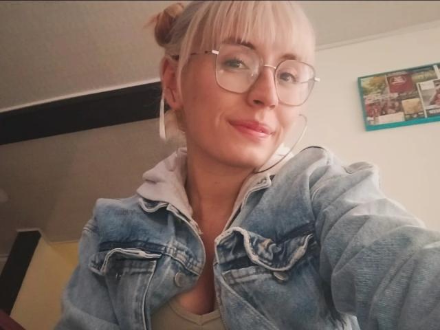 SaraleePoison heeft blond haar in een knot, draagt een bril, een denimjas en een witte hoodie, heeft oordopjes in en glimlacht lichtjes. Haar borsten zijn klein en ze is schattig..