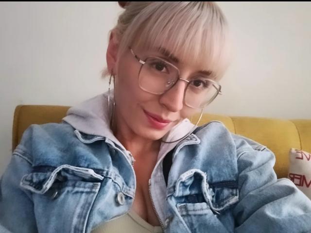 SaraleePoison with blonde hair, glasses, and a denim jacket, smiling at the camera, showing her tits slightly through the jacket, sitting on a yellow couch.