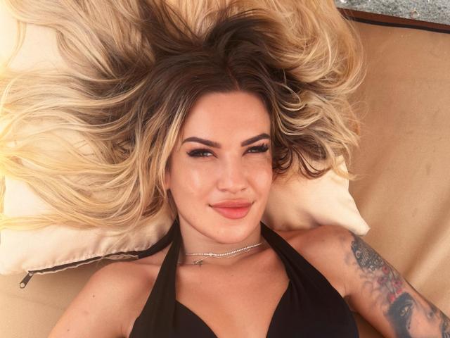 ZoeyMarie with blonde and brown ombre hair, black halter top, tattoo on right arm, lying on tan pillow, smiling at camera, wearing silver necklace.