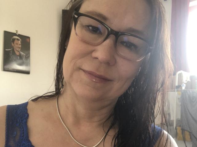 MarylouMar has wet brown hair, wears glasses, and a blue lace top with a silver necklace. Her shiny lips are slightly up, showing a subtle smile. Her tits are small.