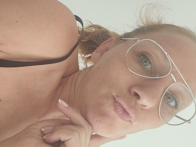 ApoloniaRed with glasses, pouty lips, and light freckles, showing off her bare shoulders and black strap, touching her chin with her finger. Boobs partially visible.