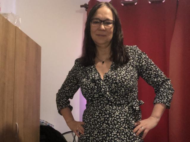 MarylouMar stands with hands on hips, wearing a black floral dress, glasses, and a necklace, in a room with a red curtain and wooden cabinet.