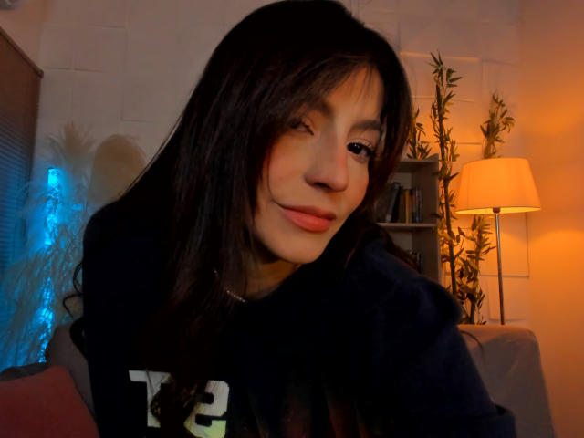 ValenRosee, winking with a slight smile, has long dark hair, wearing a black hoodie, and is in a dimly lit room with a lamp and bamboo decor.