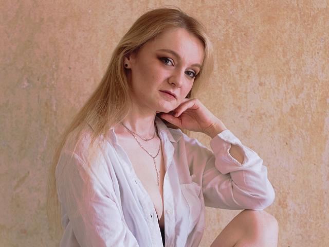 AlmaFlowers with blonde hair, wearing a white button-down shirt, exposing her small tits, sitting with one knee up, hand on chin, looking at the camera, pink wall background.