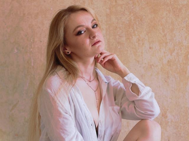 AlmaFlowers, avec ses cheveux blonds, sa chemise blanche et son collier, assise, regardant vers le haut, la main sur le menton, montrant de petites seins, un maquillage subtil, une lumière douce, un mur texturé en arrière-plan..