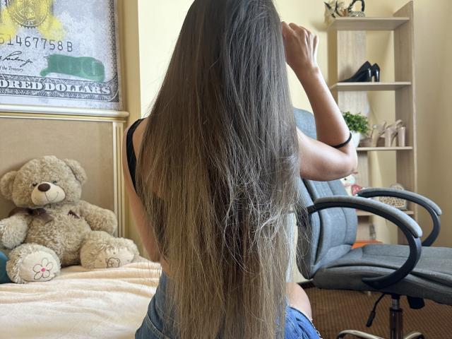 Stefanni with long brown hair, back to camera, sitting on gray chair, wearing black top, blue shorts, hand in hair, next to beige teddy bear and wooden shelves.